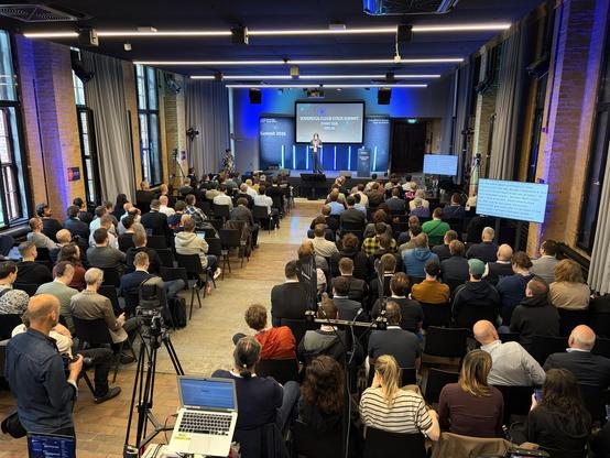 Audience attending the Sovereign Cloud Stack Summit in a spacious, modern conference room with a stage and large screens.