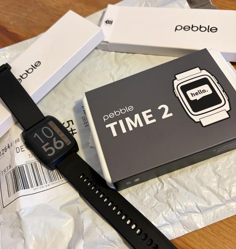 A Pebble Time 2 smartwatch displaying the time and date, with box packaging and shipping label on a crumpled paper surface.