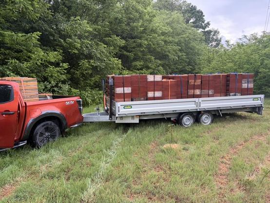 Ein roter Pickup steht auf einer grünen Wiese am Waldrand. Er zieht einen Anhänger, der mit zahlreichen braunen Bienenkisten beladen ist, auf denen jeweils Honigräume platziert sind. Die Kästen sind mit Spanngurten gesichert. Auch auf der Ladefläche des Pickups befinden sich weitere dieser gestapelten Bienenkisten mit Honigräumen.
