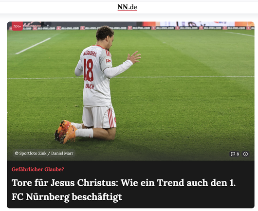The image shows a professional soccer player from behind as he kneels on a green soccer pitch. 

The player is wearing a white uniform with red details. The back of his jersey features the word NÜRNBERG printed in red capital letters, with the number 18 below it. He has dark, curly hair and is kneeling on both knees with his lower legs flat on the grass. His feet are angled back, showing the bottoms of his bright orange and black soccer cleats. 

His posture is solemn. His head is slightly bowe…