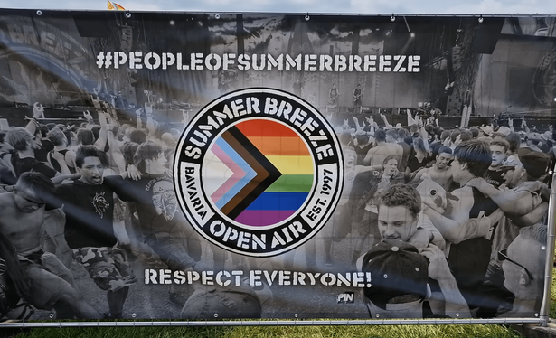 Großes Banner am Zaun eines Festivalgeländes. Hintergrund: schwarzweißes Foto einer feiernden Menge vor der Bühne. Oben in weißer Schablonenschrift "#PEOPLEOFSUMMERBREEZE", unten "RESPECT EVERYONE!". In der Mitte das runde Summer-Breeze-Logo mit dem Schriftzug "Summer Breeze, Bavaria Open Air, est. 1997". Die Innenfläche zeigt die Progress-Pride-Flagge: Regenbogen mit vorgelagertem Pfeil in Hellblau, Rosa, Weiß, Braun und Schwarz.