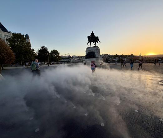 People walking around a large plaza at sunset with a statue of a person on horseback in the background, steam rising from the ground.