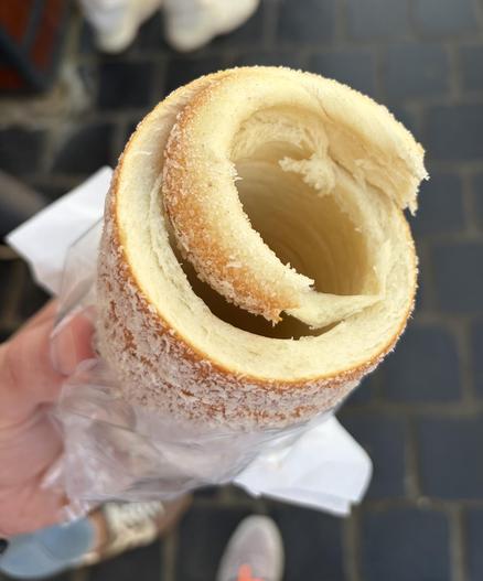 A hand holding a partially eaten Swiss roll cake with outer coating of coconut flakes, on a clear plastic wrap, with a brick pavement background.