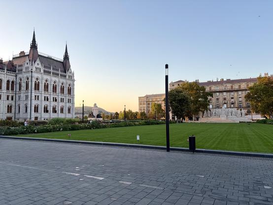 A historic white stone building with pointed towers on the left, a green lawn with pathways in the center, and residential buildings with trees on the right.