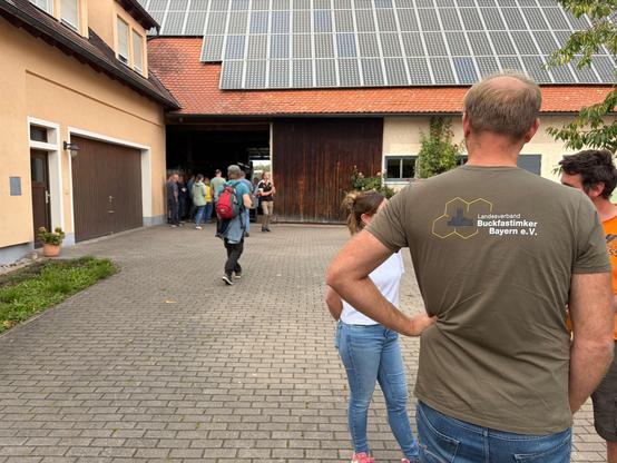 Hofeinfahrt eines Bauernhofs mit großer Photovoltaikanlage auf dem Dach. Eine Gruppe von Menschen steht vor dem offenen Scheunentor, weitere gehen darauf zu. Im Vordergrund von hinten ein Mann in olivgrünem T-Shirt mit dem Logo „Landesverband Buckfastimker Bayern e.V.“ (gelbe Waben mit Burgmotiv), daneben zwei weitere Personen.