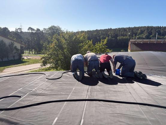 Vier Männer knien bei Sonnenschein auf einer großen schwarzen Biogasspeichermembran und arbeiten am Rand, einer hält einen blauen Eimer. Über die Membran läuft ein schwarzer Druckluftschlauch. Im Hintergrund Hallen, Wiesen und bewaldete Hügel unter blauem Himmel.