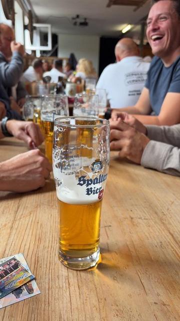 The photo shows a half-full glass of Spalter beer in focus on a light wooden table. It is surrounded by a group of people socialising, their arms and hands resting on the table. In the background, people are laughing and more glasses can be seen. The scene feels relaxed and lively.